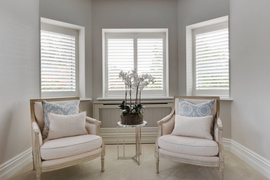 Bay window with plantation shutters, two patterned armchairs and an orchid on a glass side table