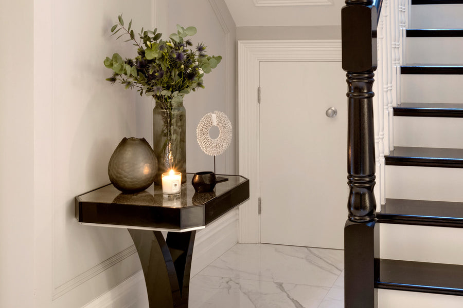 Dark timber console with a vase of foliage and a lit candle beside a black and white staircase