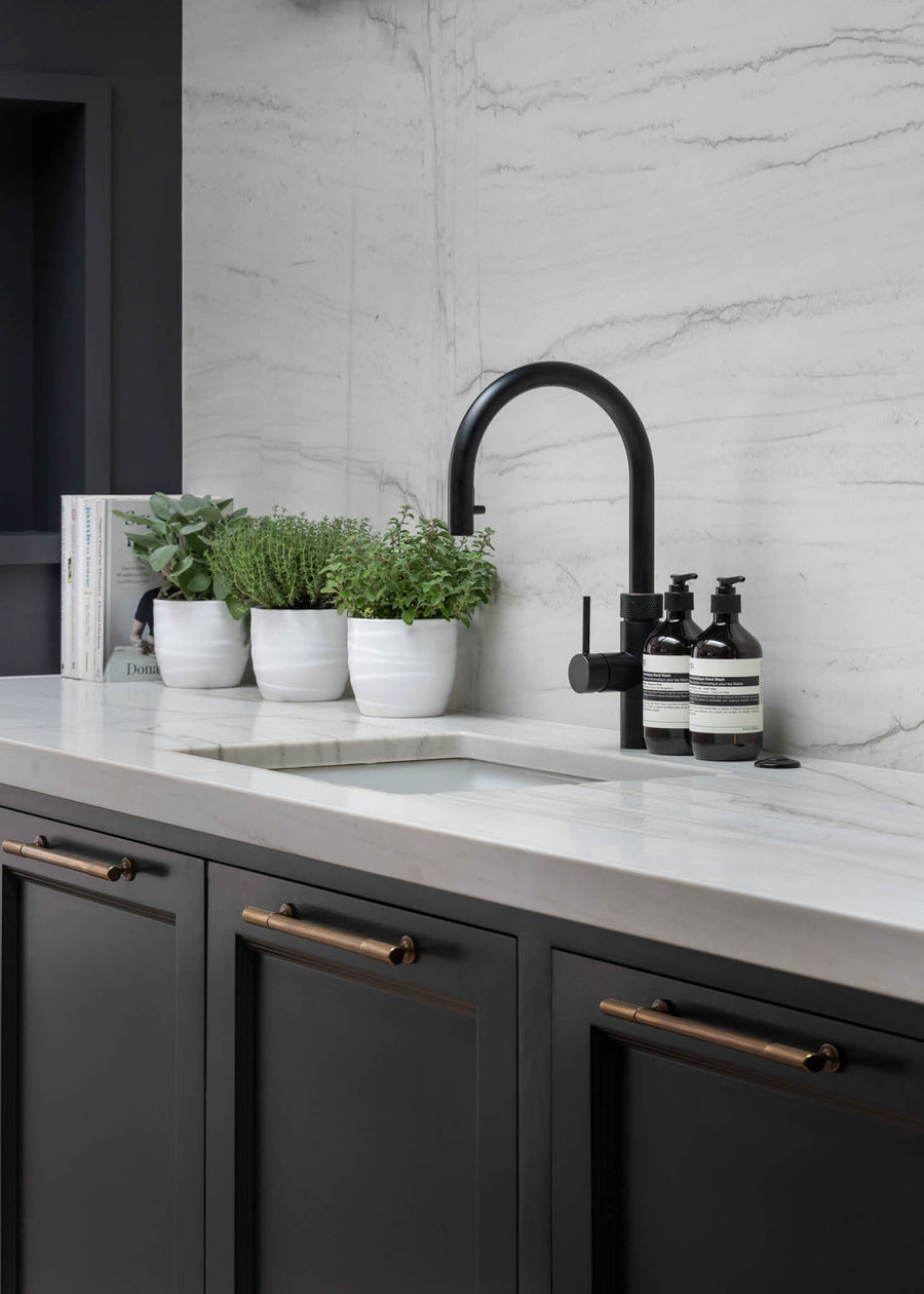 Marble splashback with a matt black tap, potted herbs and dark cabinetry with brass handles