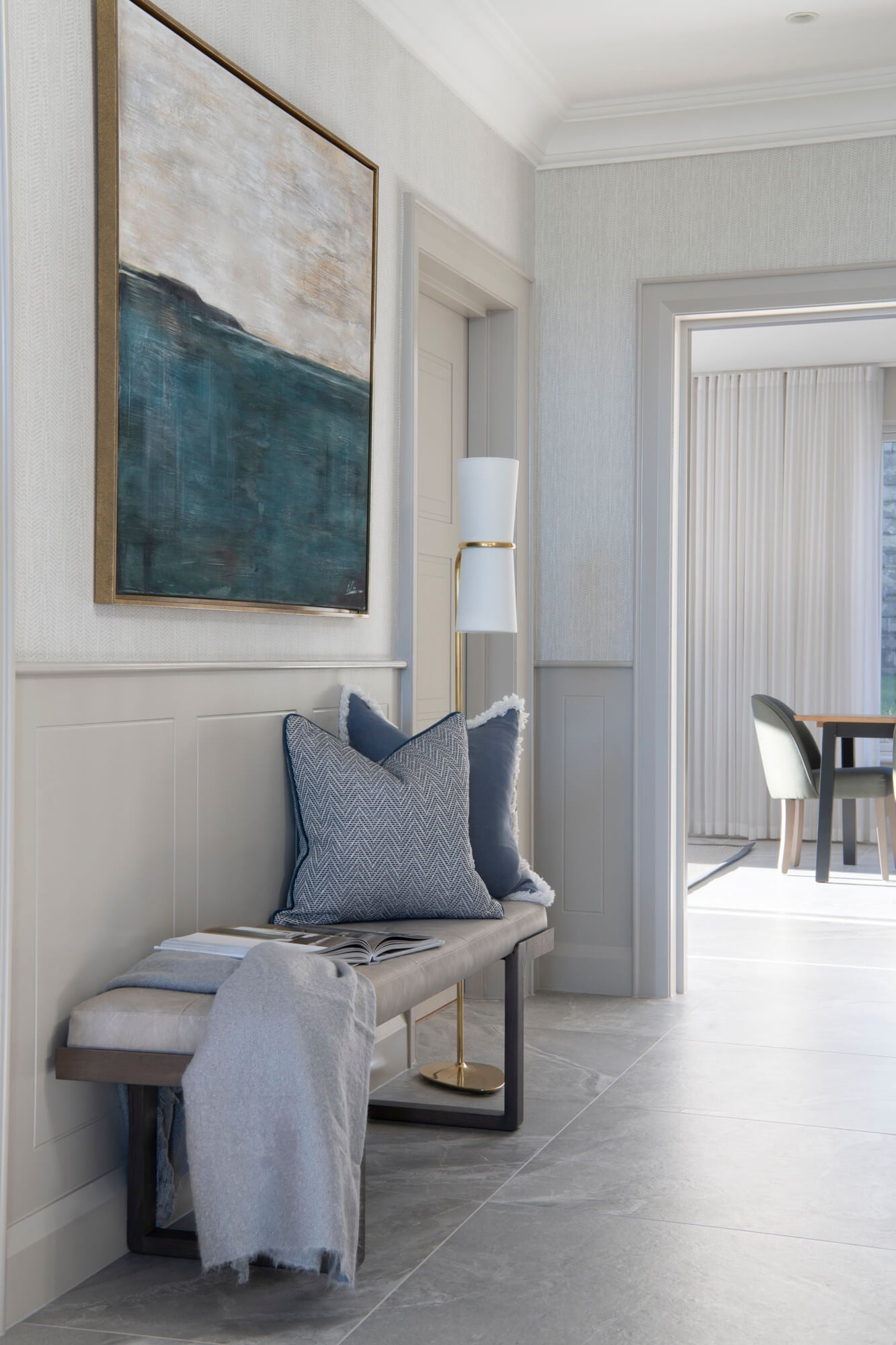Entrance hall designed by Sarah Louise Interiors: an upholstered bench with cushions and a throw beneath a large framed abstract seascape, brass floor lamp alongside.