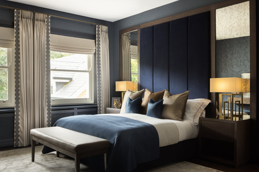 Navy panelled bedroom with a blue velvet bed, upholstered bench, brass lamps and roman blinds either side of the window