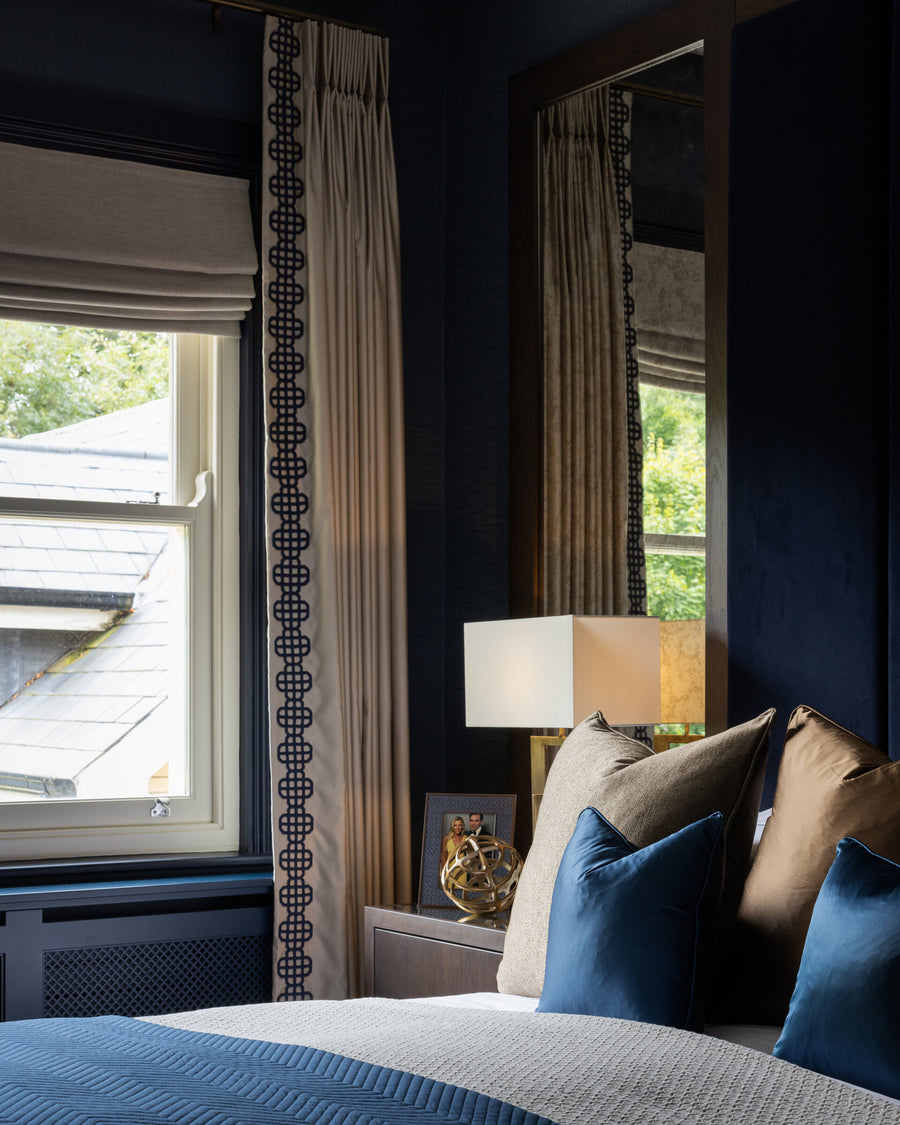 Bedroom window with braid-trimmed curtains, a tall mirror and blue and bronze cushions on the bed