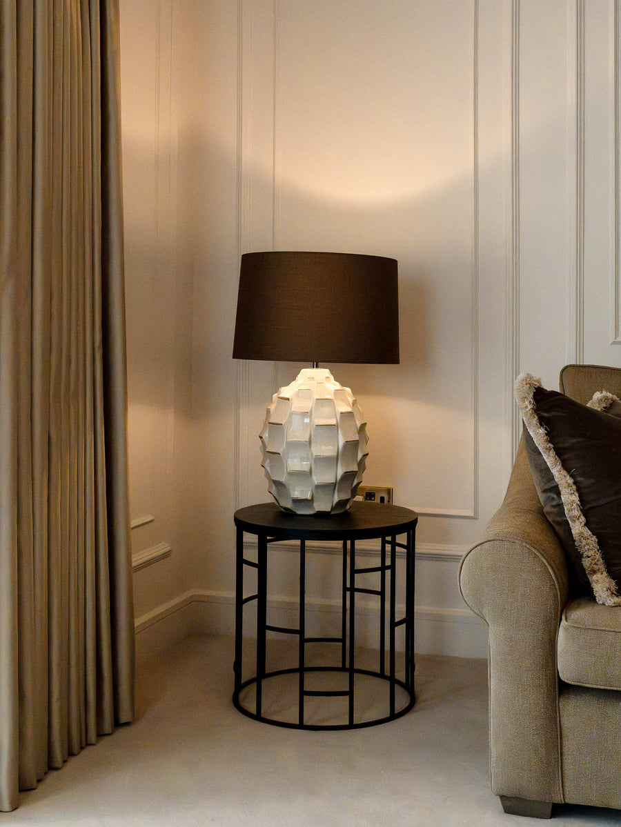 Sculptural white table lamp with a bronze shade on a slim black side table beside floor-length curtains
